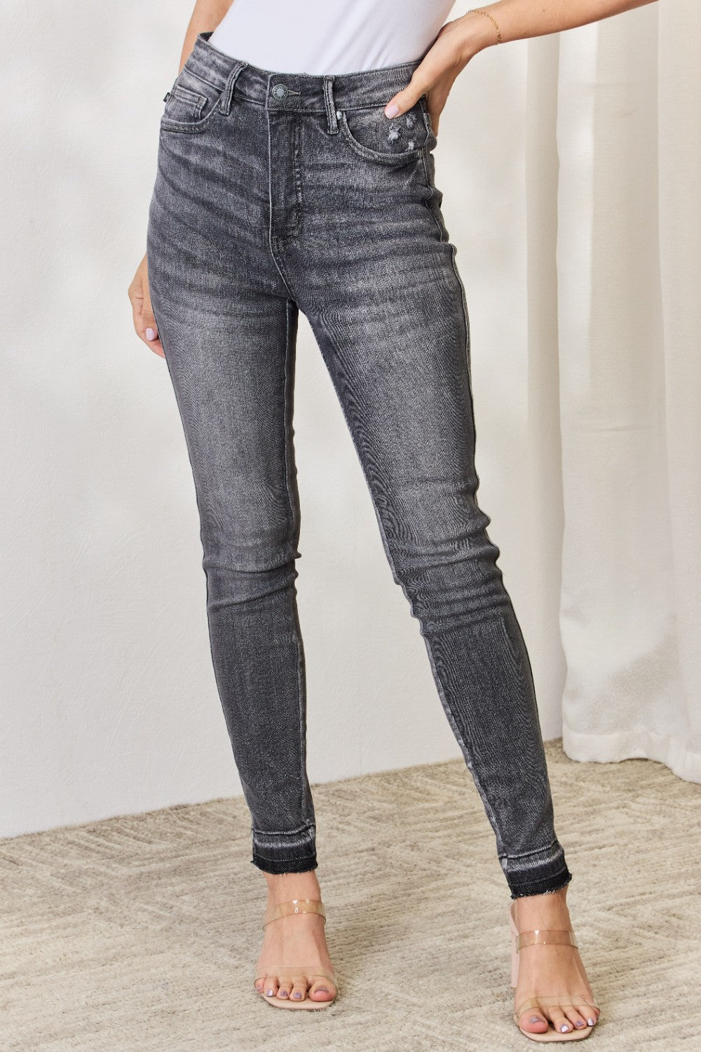 A person wearing the Judy Blue Full Size High Waist Tummy Control Release Hem Skinny Jeans paired with nude heels stands against a light background.