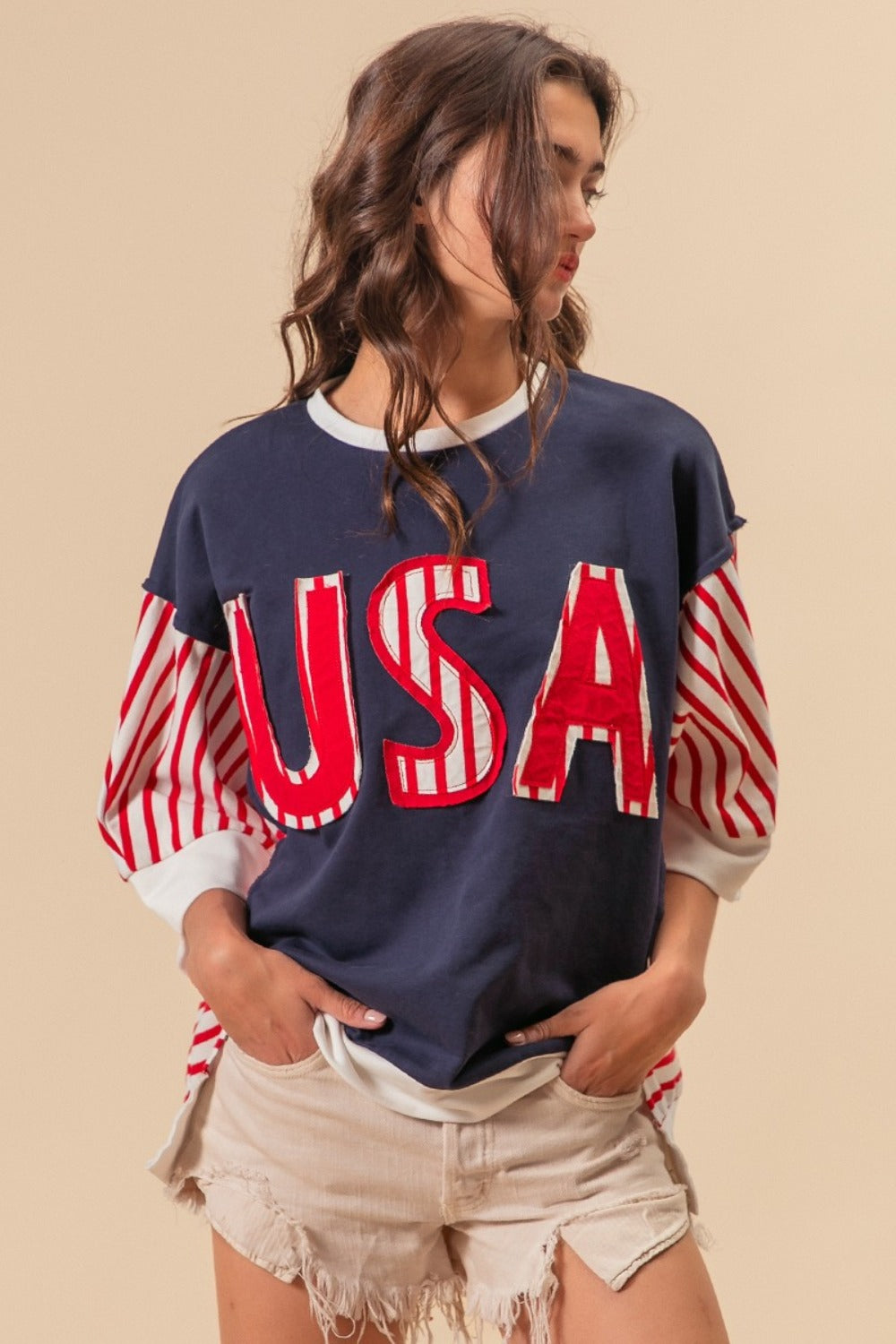 A person dressed in a “BiBi USA Letter Patchwork Contrast Short Sleeve T-Shirt” featuring blue, red, and white colors with striped sleeves, paired with beige shorts, poses against a beige background, proudly displaying their American spirit.