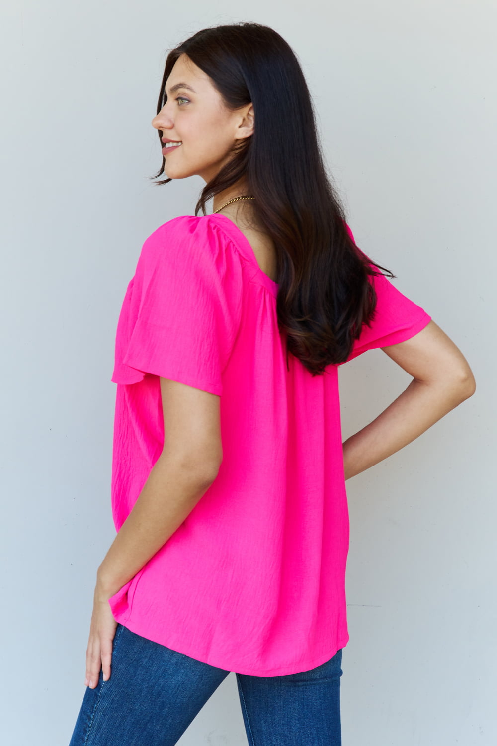 A person wearing the Ninexis Keep Me Close Square Neck Short Sleeve Blouse in Fuchsia paired with blue jeans smiles while posing against a plain background.