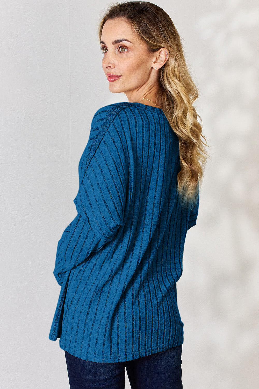 A woman with long, wavy blonde hair wears a slightly stretchy, textured Basic Bae Full Size Ribbed Half Button Long Sleeve T-Shirt in pink, standing against a neutral backdrop.