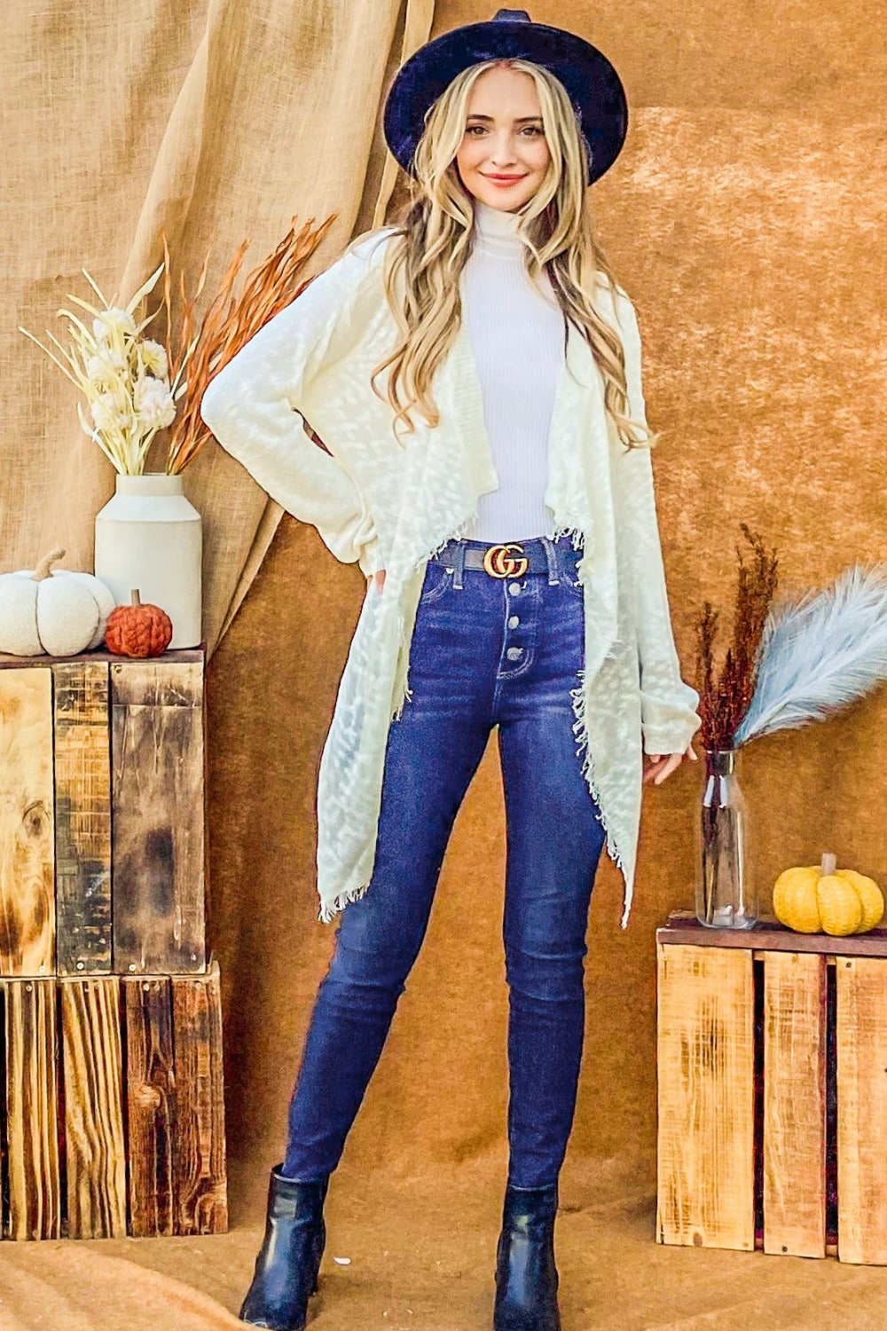 A person with long blonde hair wears a black hat, the And The Why Textured Knit Fringe Hem Cardigan in white, and blue jeans, standing against a brown backdrop near a wooden table with a vase of dried flowers and a small pumpkin.