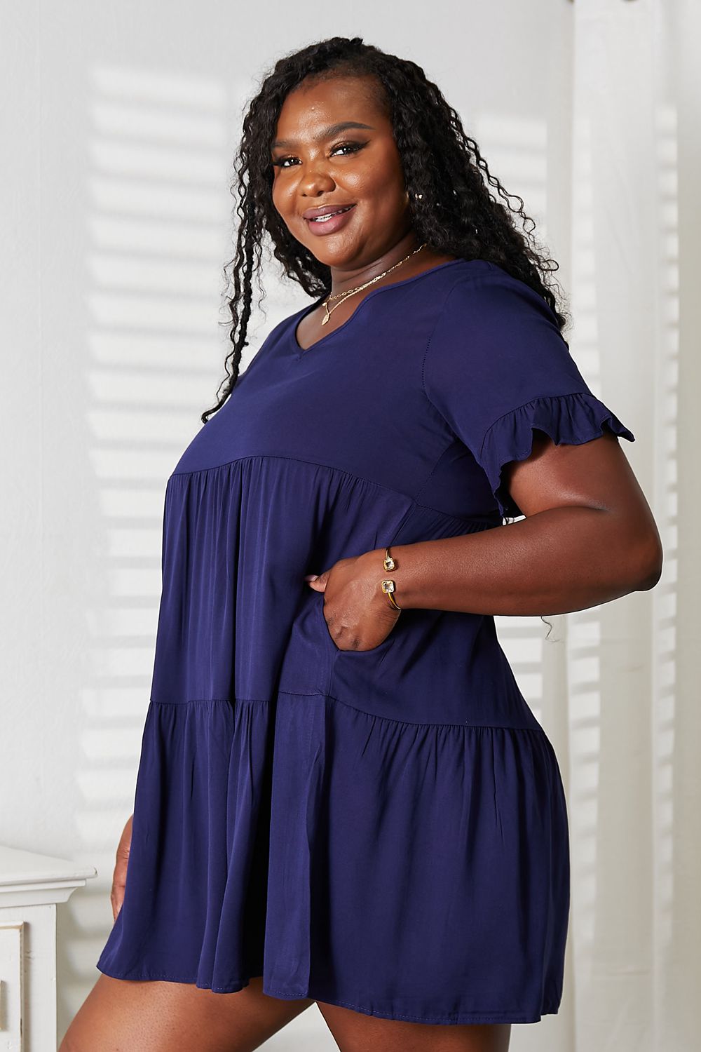 A woman stands indoors wearing the Mandy V-Neck Flounce Sleeve Tiered Dress in navy blue. She has one hand on her hip and is smiling.