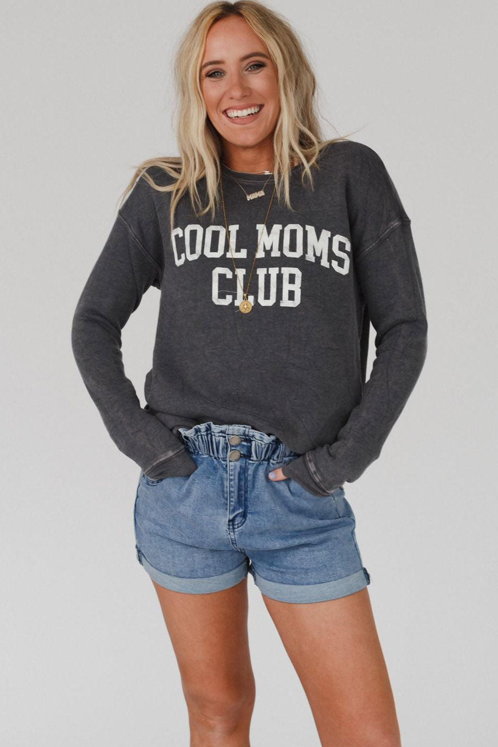 A person with long blond hair wearing a gray COOL MOMS CLUB drop shoulder sweatshirt and relaxed-fit blue jeans is seen from the back, standing near a pumpkin and a window.