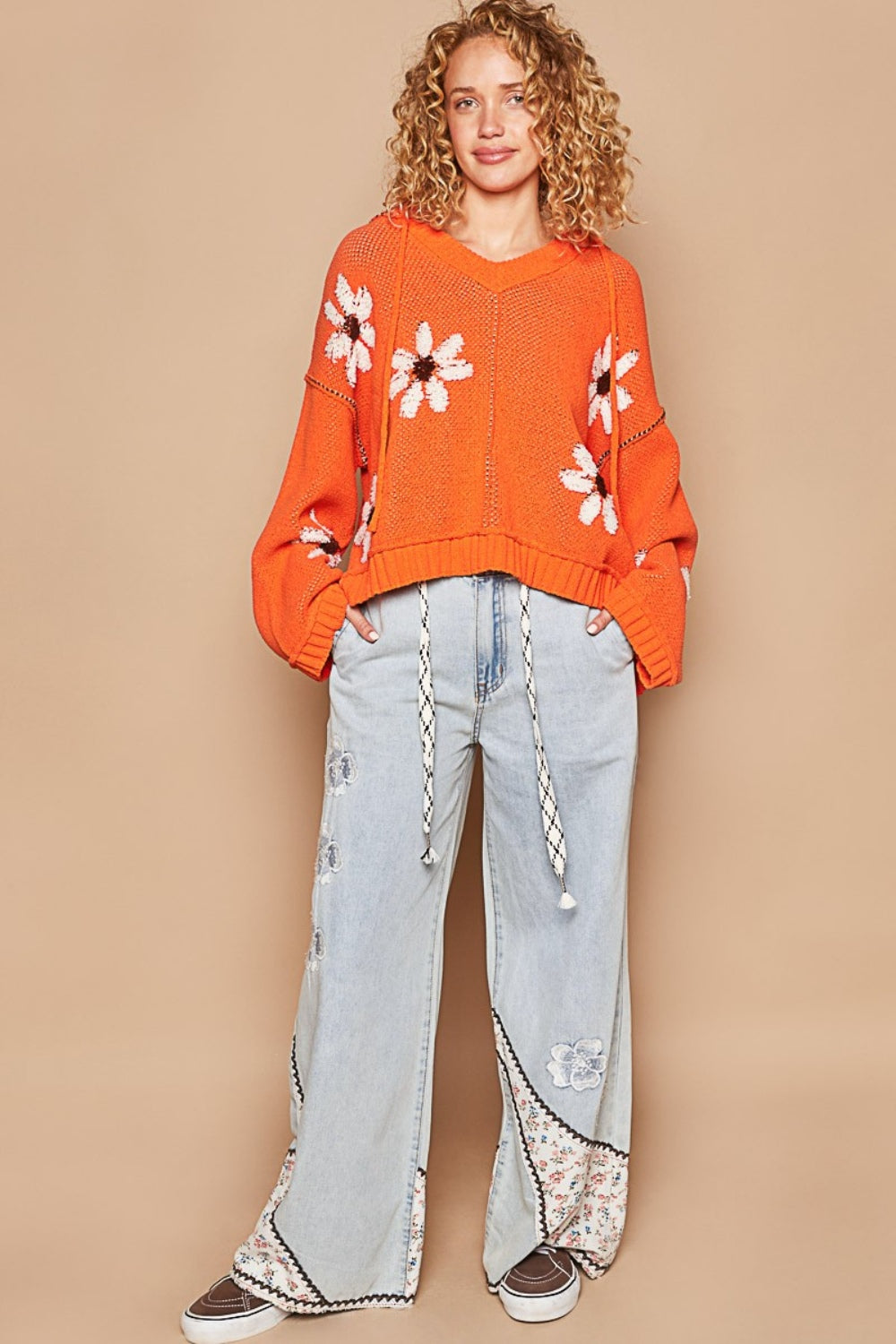 A person with curly blond hair is wearing the POL Floral Pattern Hooded High-Low Sweater in orange, featuring white and brown flower patterns against a solid beige background, paired with light blue jeans.