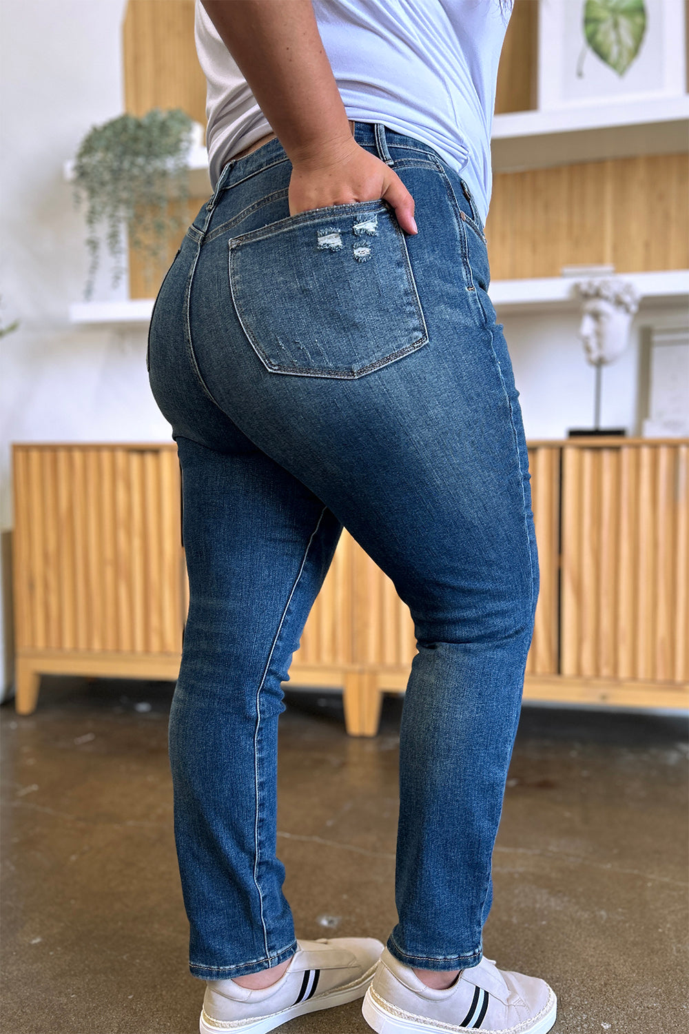 Person wearing Judy Blue Full Size Tummy Control High Waist Slim Jeans in blue and a white shirt, with their hands in their pockets. They are standing indoors on a polished concrete floor with a wooden cabinet and plant in the background.