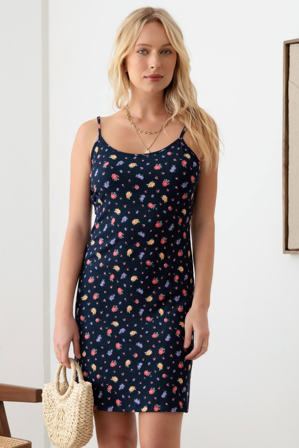 A woman with long blonde hair is wearing a Gilli Floral Ribbed Tie-Back Cami Mini Dress. She is holding a woven straw handbag and standing in a minimalist room.