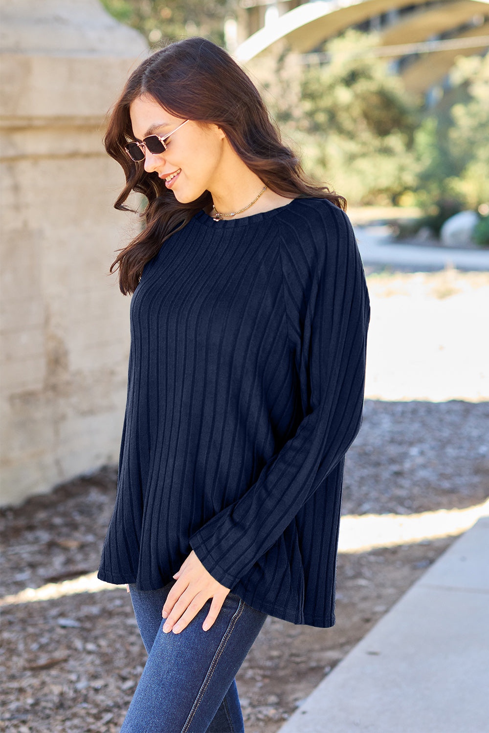 A woman with long brown hair wearing sunglasses and a Basic Bae full-size, slightly stretchy ribbed round neck long sleeve knit top in red, paired with blue jeans, is standing outdoors.