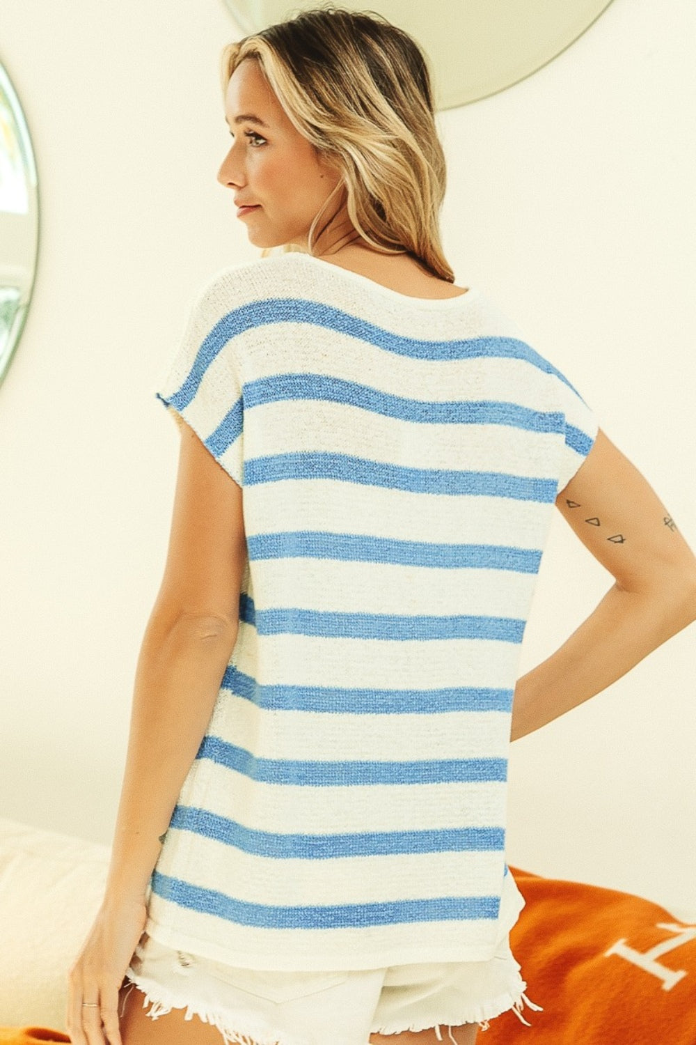 A person with long, wavy hair is smiling while wearing the versatile and chic BiBi Striped Round Neck Short Sleeve Knit Top in blue and white, paired with white shorts. They are standing in a brightly lit room with natural light streaming through a window.