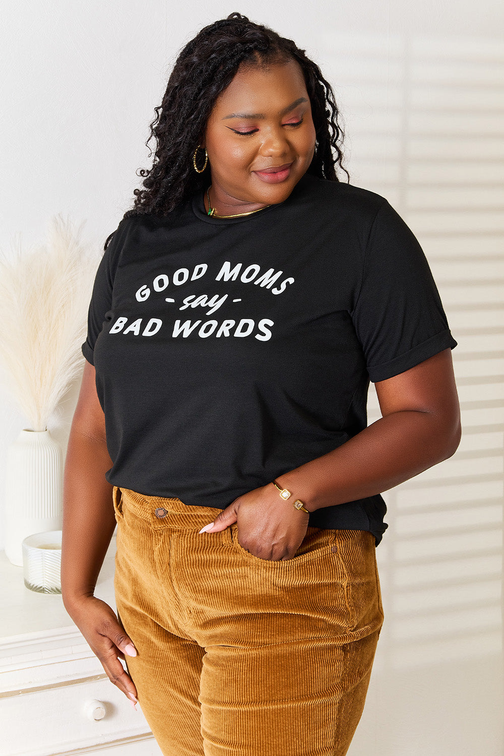 A woman stands in a room with a white wall, where light filters through the blinds, subtly showcasing her rebellious side while wearing the Simply Love GOOD MOMS SAY BAD WORDS Graphic Tee.