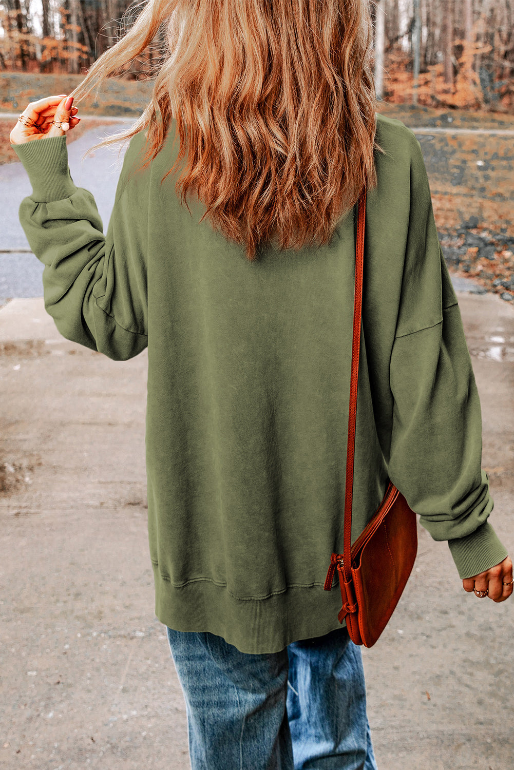 A person with wavy, light brown hair, wearing a Green Drop Shoulder Ribbed Trim Oversized Sweatshirt made of skin-friendly fabrics, blue jeans, and holding a brown shoulder bag is walking outdoors on a concrete path.