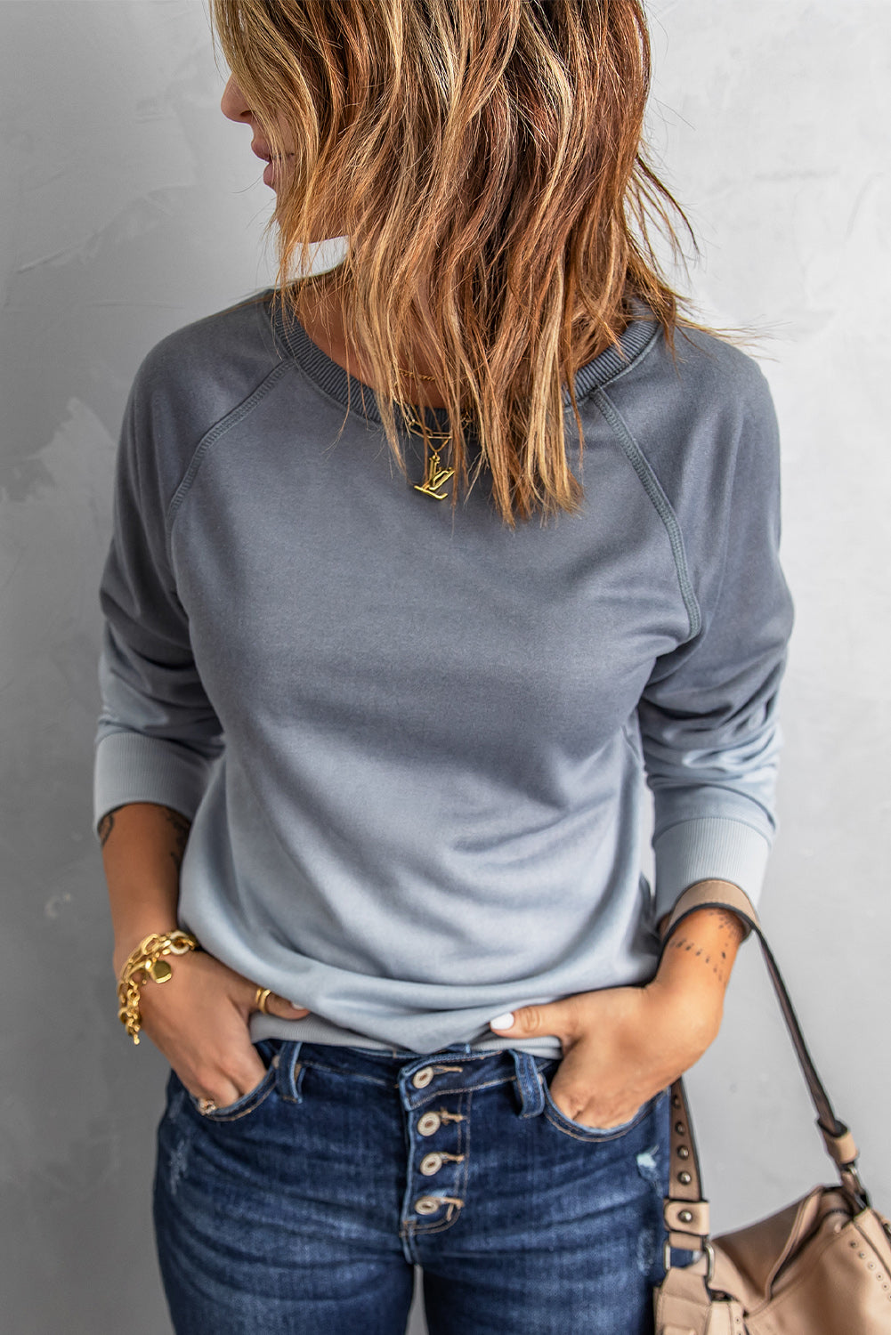 A person with wavy hair stands facing a gray wall, wearing a Gray Ombre Crewneck Long Sleeve Sweatshirt and blue jeans, with one hand on their hip.