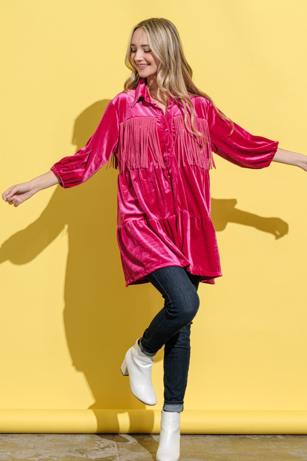 A woman stands against a bright yellow background, smiling and wearing an elegant statement fringed, bright pink And The Why Fringe Detailed Velvet Shirt Dress.