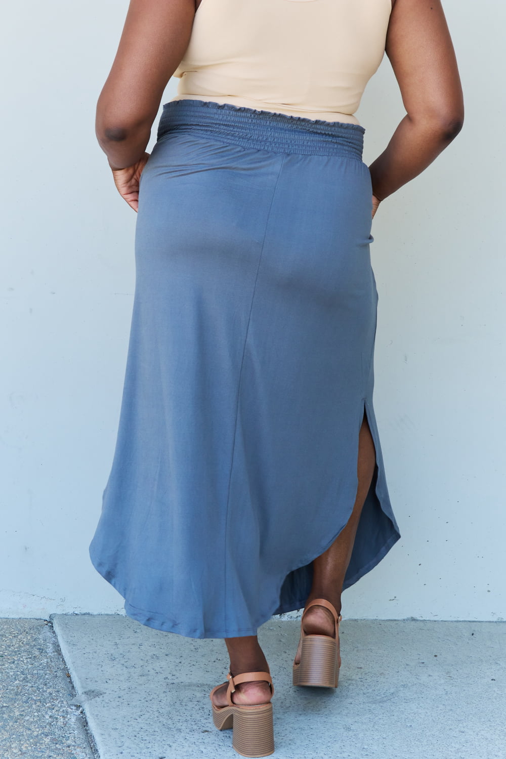 A person wearing a white sleeveless top and the Doublju Comfort Princess Full Size High Waist Scoop Hem Maxi Skirt in Charcoal stands against a plain wall. They are also wearing black sandals.