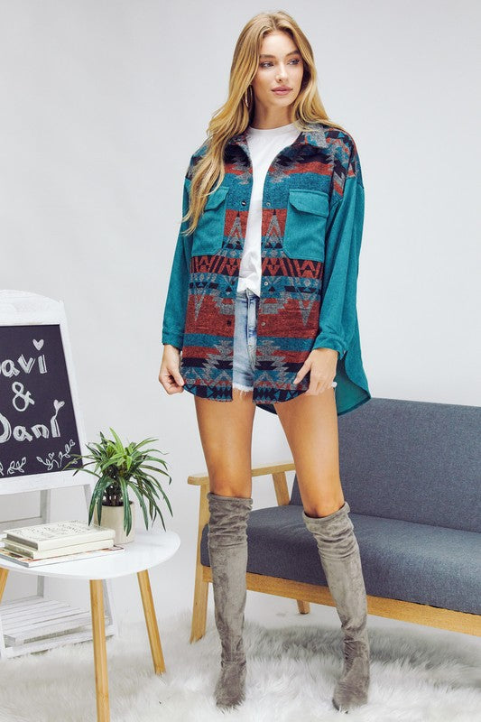 A woman with long blonde hair wears a blue and gray patterned shirt over a white crop top and dark jeans, standing indoors near a window, her casual **Printed Button Down Long Sleeve Jacket** draped casually over her shoulders.