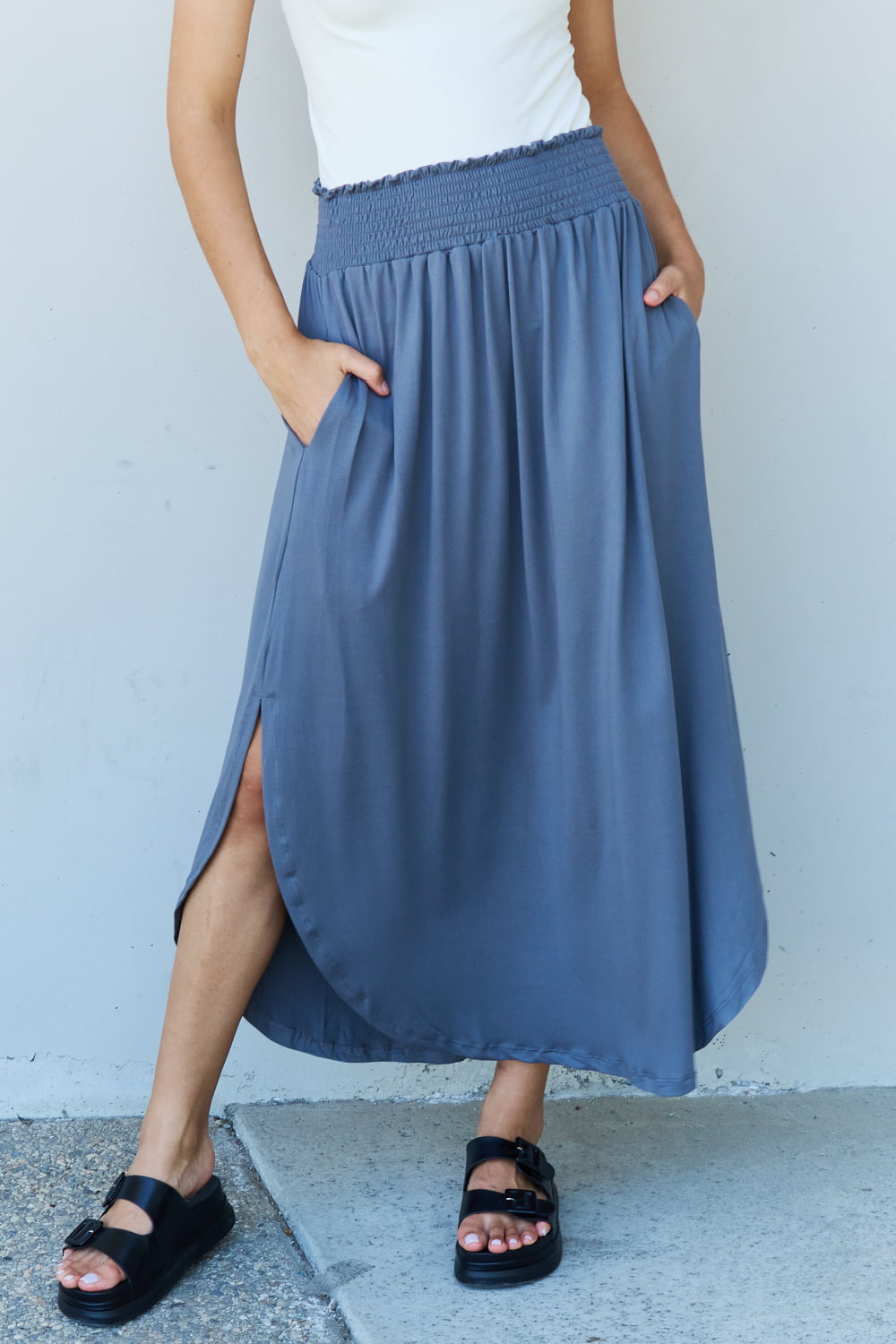 A person wearing a white sleeveless top and the Doublju Comfort Princess Full Size High Waist Scoop Hem Maxi Skirt in Charcoal stands against a plain wall. They are also wearing black sandals.