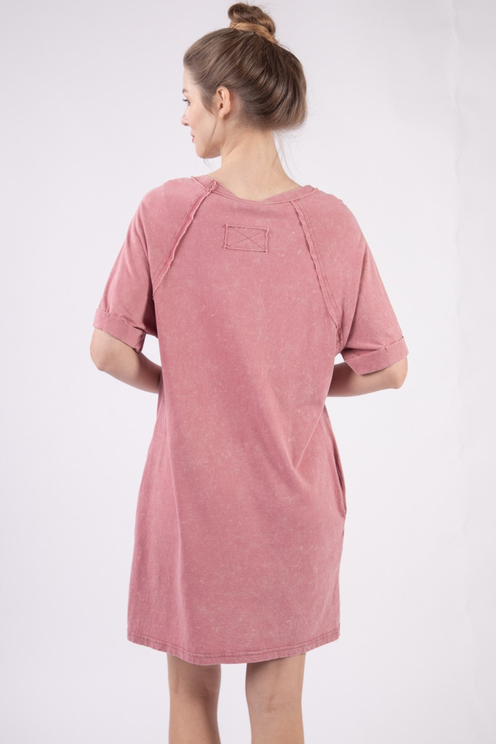 Wearing the VERY J Washed Round Neck Mini Tee Dress in pink, a person stands sideways against a plain background, effortlessly showcasing their casual wardrobe style.