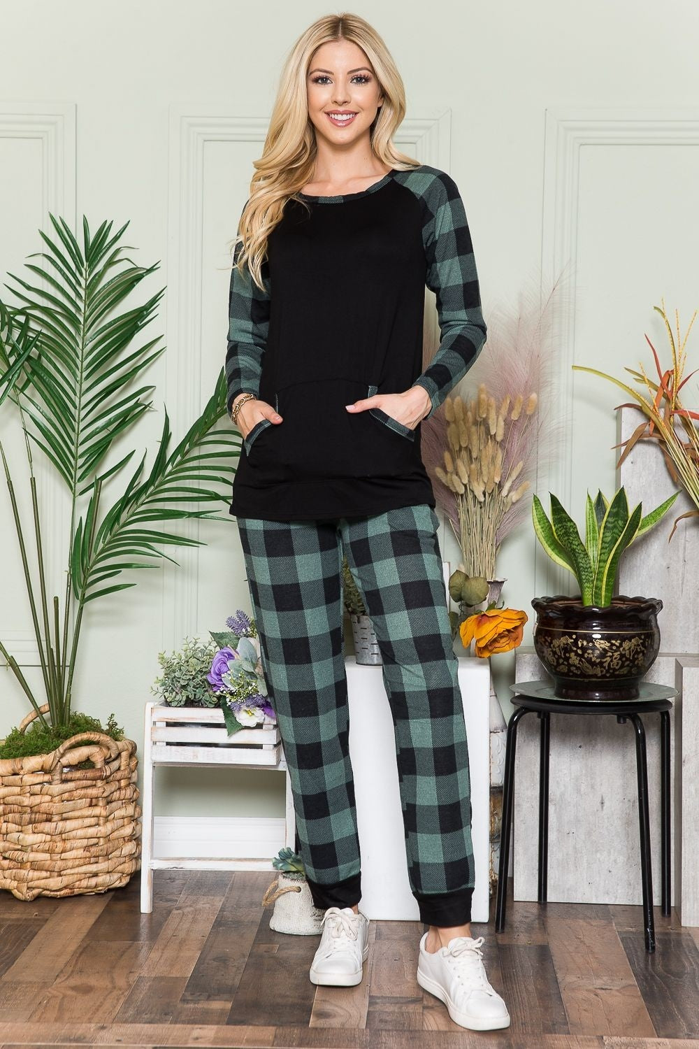 An individual wearing a Celeste Plaid Long Sleeve T-Shirt with Pockets stands indoors on a wooden floor, next to potted plants and a wicker basket.