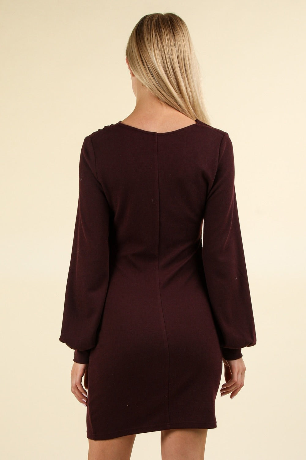 A person with long blonde hair is wearing a VERY J Ruched Detail Bodycon Mini Dress in burgundy, standing against a plain background. The figure-hugging silhouette of this dress makes it perfect for special occasions.