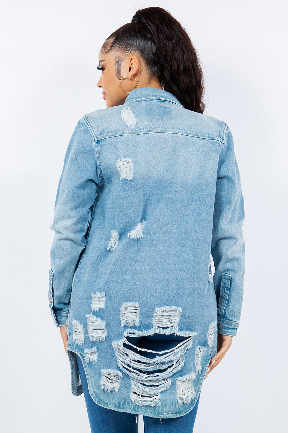 A woman with long hair is wearing an American Bazi Distressed Button Up Long Sleeve Denim Jacket over a white crop top and blue jeans. She is facing forward and standing against a plain white background.