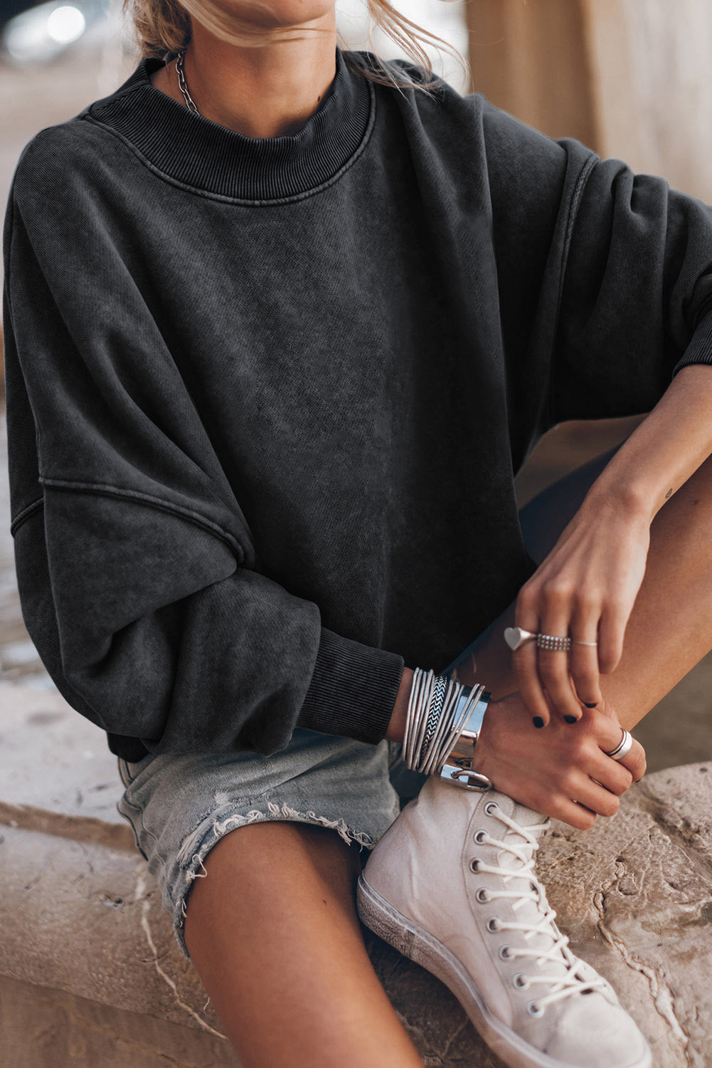 Person wearing a black drop shoulder crew neck pullover sweatshirt with rolled-up sleeves and light blue jeans, standing with one hand in their pocket, showing multiple bracelets on their wrist.