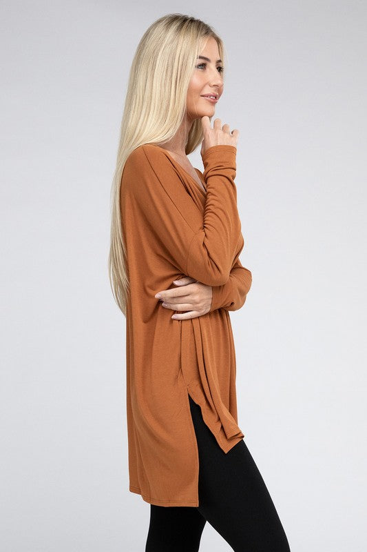 A woman with long, blonde hair is wearing a loose-fitting, brown Dolman Long Sleeve V-Neck Side Slit Hi-Low Hem Top and black pants. She poses with one hand on her hip and a neutral facial expression against a light grey background.