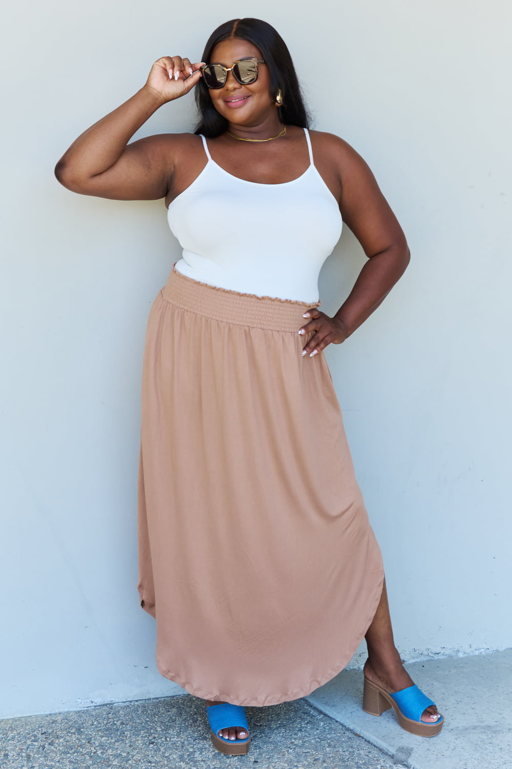 A person stands against a wall, wearing a white sleeveless top, the Doublju Comfort Princess Full Size High Waist Scoop Hem Maxi Skirt in Tan, and blue platform sandals. The right hand rests on the side of the skirt.