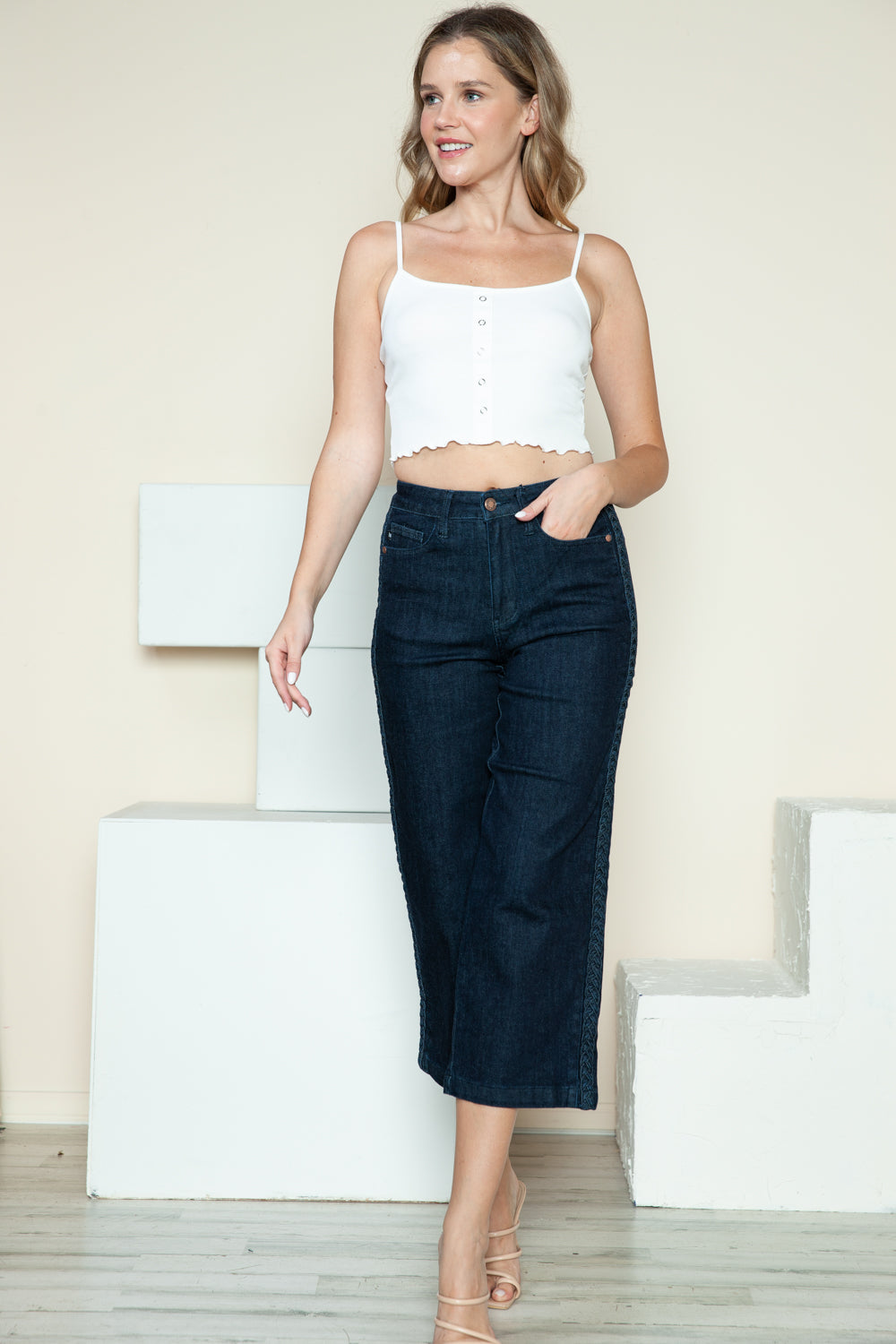 Person wearing Judy Blue Full Size Side Seam Braid Detail Crop Wide Leg Jeans and beige strappy heels, standing on a wooden floor with a white background.