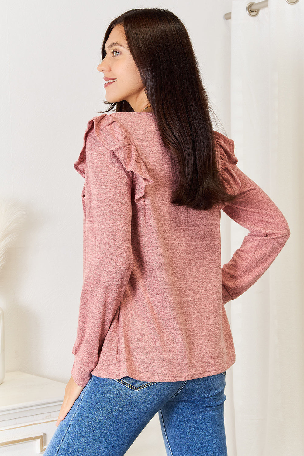 A woman wearing a Double Take Square Neck Ruffle Shoulder Long Sleeve T-Shirt in pink and blue jeans stands with one hand in her pocket, smiling. She has long curly hair and wears a necklace and bracelet. She stands in a room with white decor.