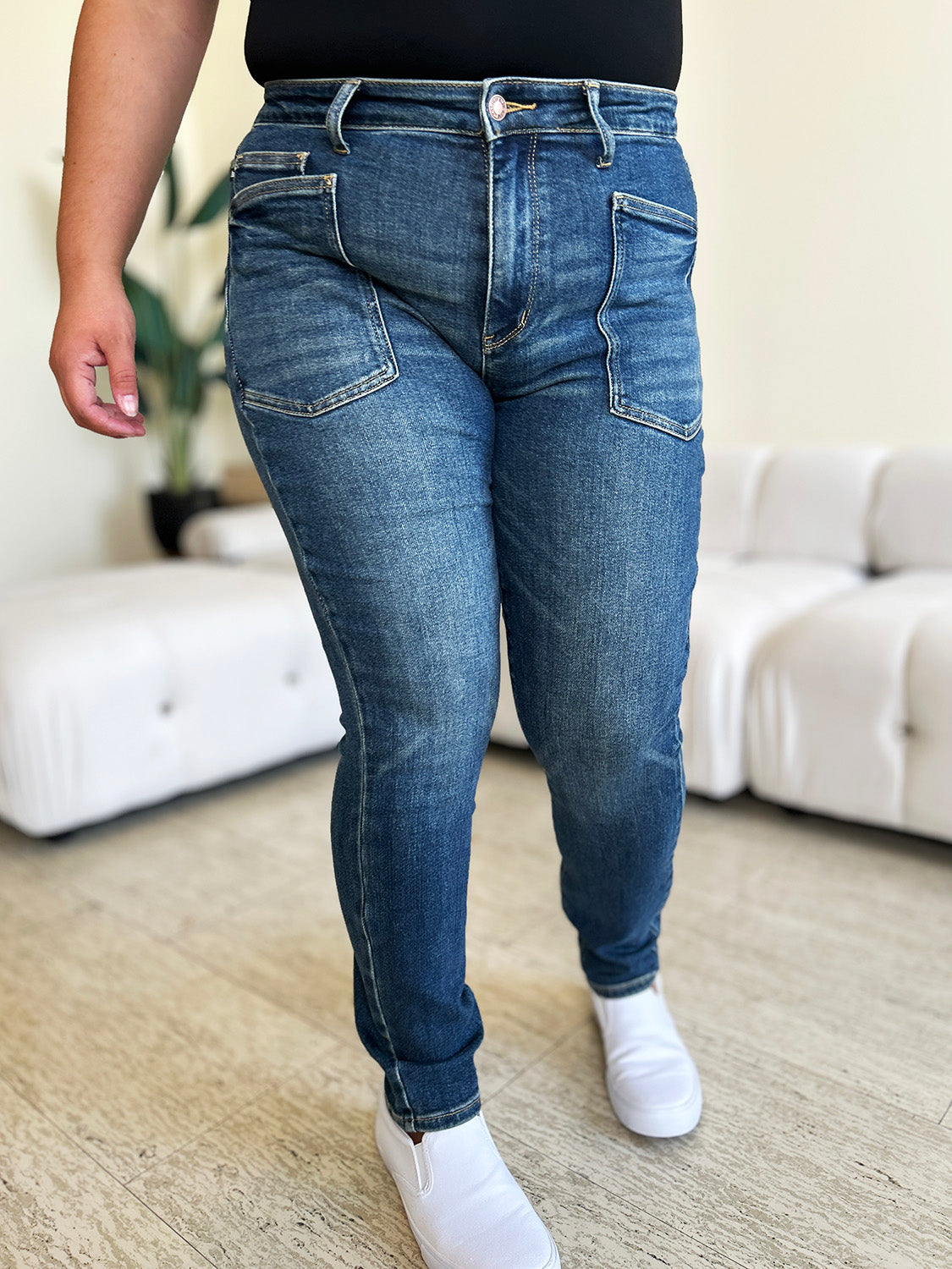 A person wearing Judy Blue Full Size High Waist Skinny Jeans made of stretchy denim fabric stands in a room with light-colored furniture and wooden flooring, paired perfectly with white slip-on shoes.