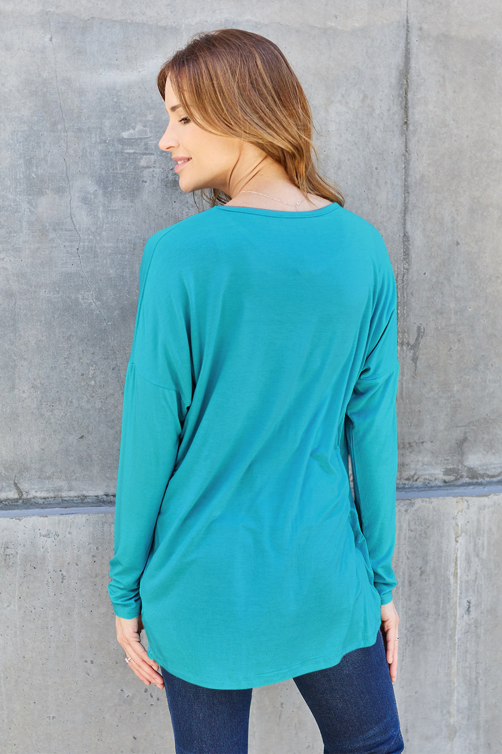 A woman with shoulder-length hair wearing a Basic Bae Full Size Round Neck Dropped Shoulder T-Shirt in green and a pair of jeans, exuding a basic style, stands against a concrete wall. She's holding an imported brown woven backpack on her shoulder while touching her hair with her hand.