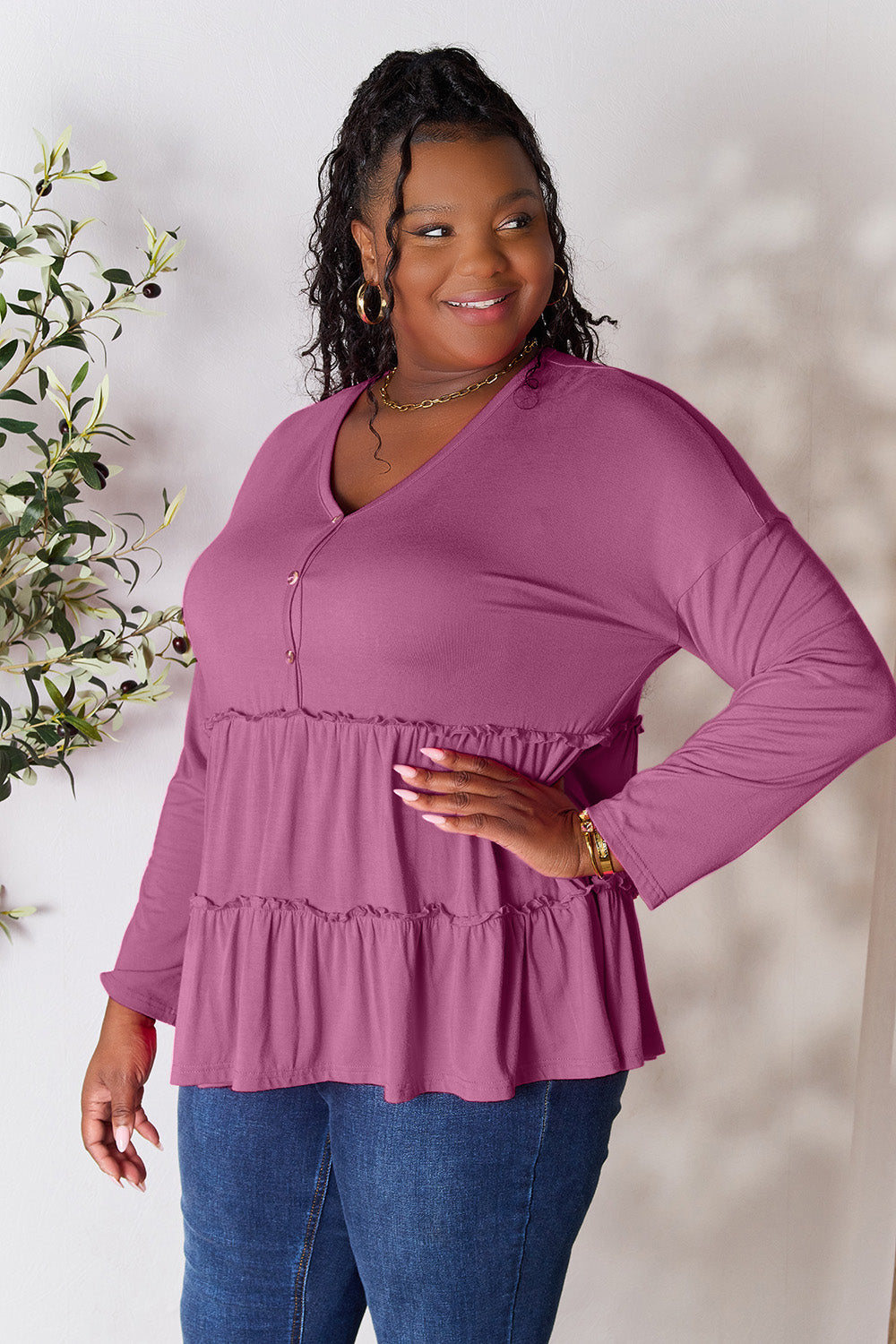 Person stands indoors near a plant, wearing the Double Take Half Button Long Sleeve Ruffle Hem Blouse in gray and blue jeans, smiling while looking to the side. The slightly stretchy fabric of the blouse adds comfort while its buttoned design offers a touch of elegance.