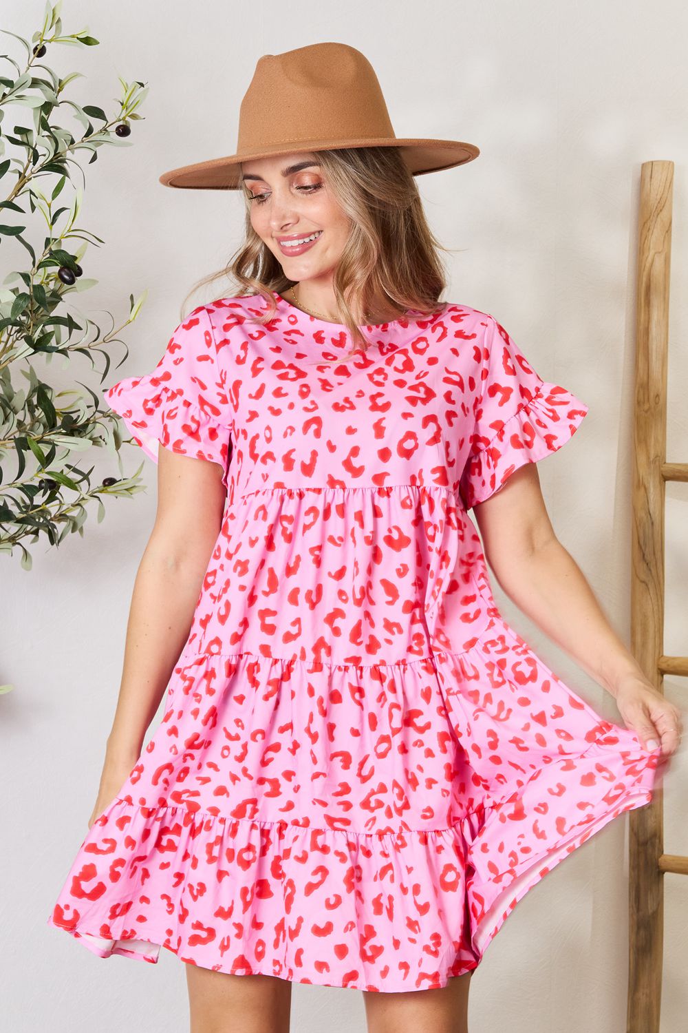 A woman wearing the Double Take Short Flounce Sleeve Tiered Dress in pink and red leopard print, complete with a ruffled hem and short flounce sleeves, smiles as she adjusts her brown hat. She stands near a plant, embodying the perfect boho-chic look.