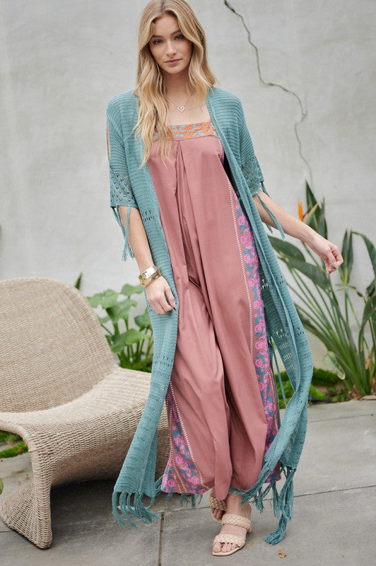 A woman in a green floral dress and a Solid Long Cardigan With Fringe exudes boho-chic vibes as she strolls on a patio, surrounded by outdoor furniture and lush plants.