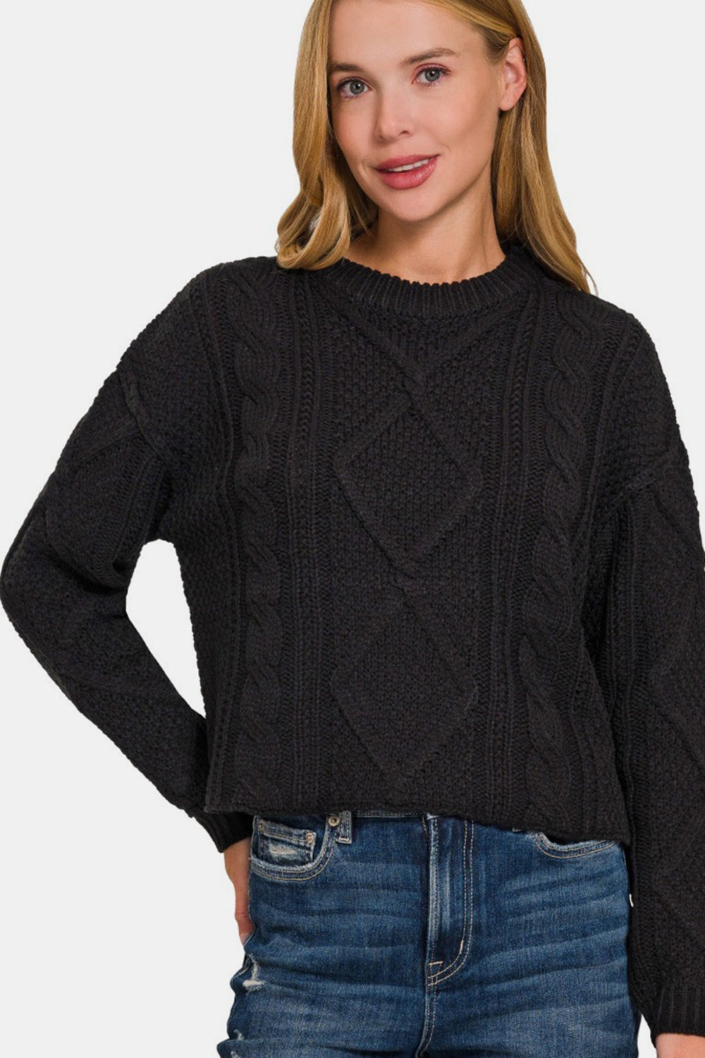 A person wearing a Zenana Cropped High Low Cable Sweater with Side Slits in black and blue jeans stands against a light gray background.