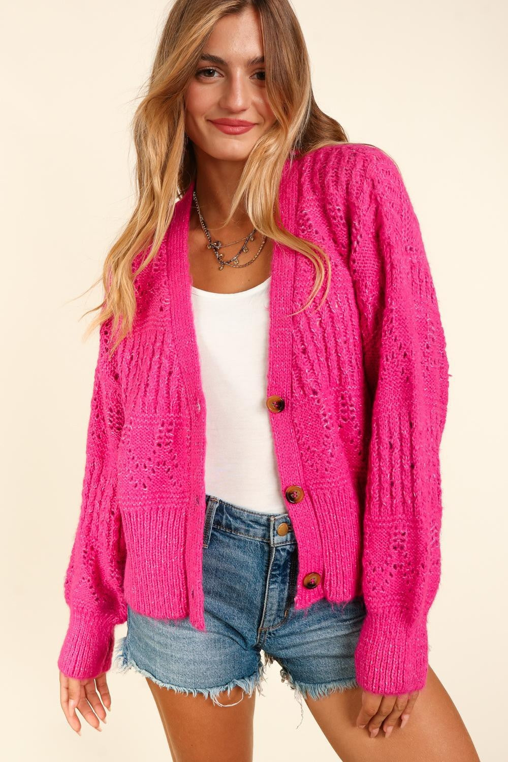 A person with long hair wears the trendy, bright pink Haptics Full Size Button Down Crop Long Sleeve Cardigan over a white top and denim shorts. They hold one side of the cardigan's collar and smile at the camera, showcasing their stylish and versatile layering.