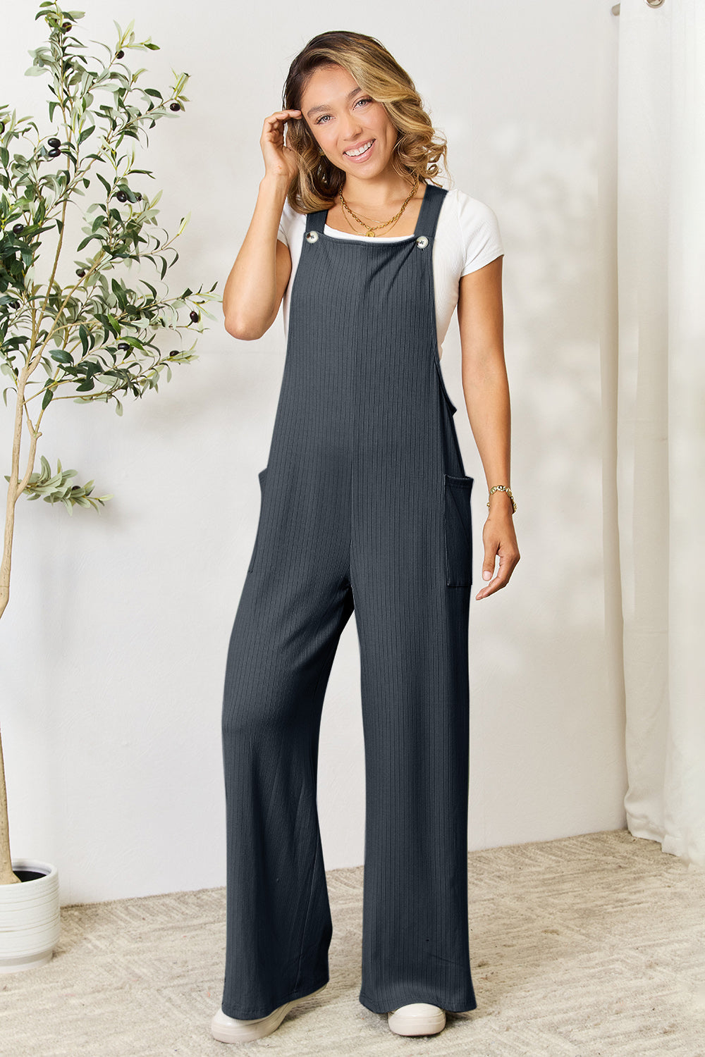 A smiling person wearing the "Double Take Full Size Wide Strap Overall with Pockets," known for its basic style and size-inclusive fit, stands indoors next to a potted plant.