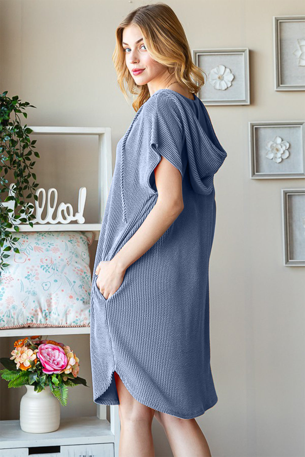 A woman wearing the Heimish Full Size Ribbed Short Sleeve Hooded Dress in a relaxed blue fit stands next to a window, touching her hair. A shelf with decorative items is visible in the background.