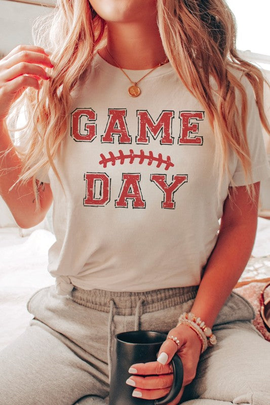 A young woman with long brown hair is wearing a 100% cotton GAME DAY Graphic Tee, featuring a football graphic and "Game Day" text, as she stands against a neutral background.