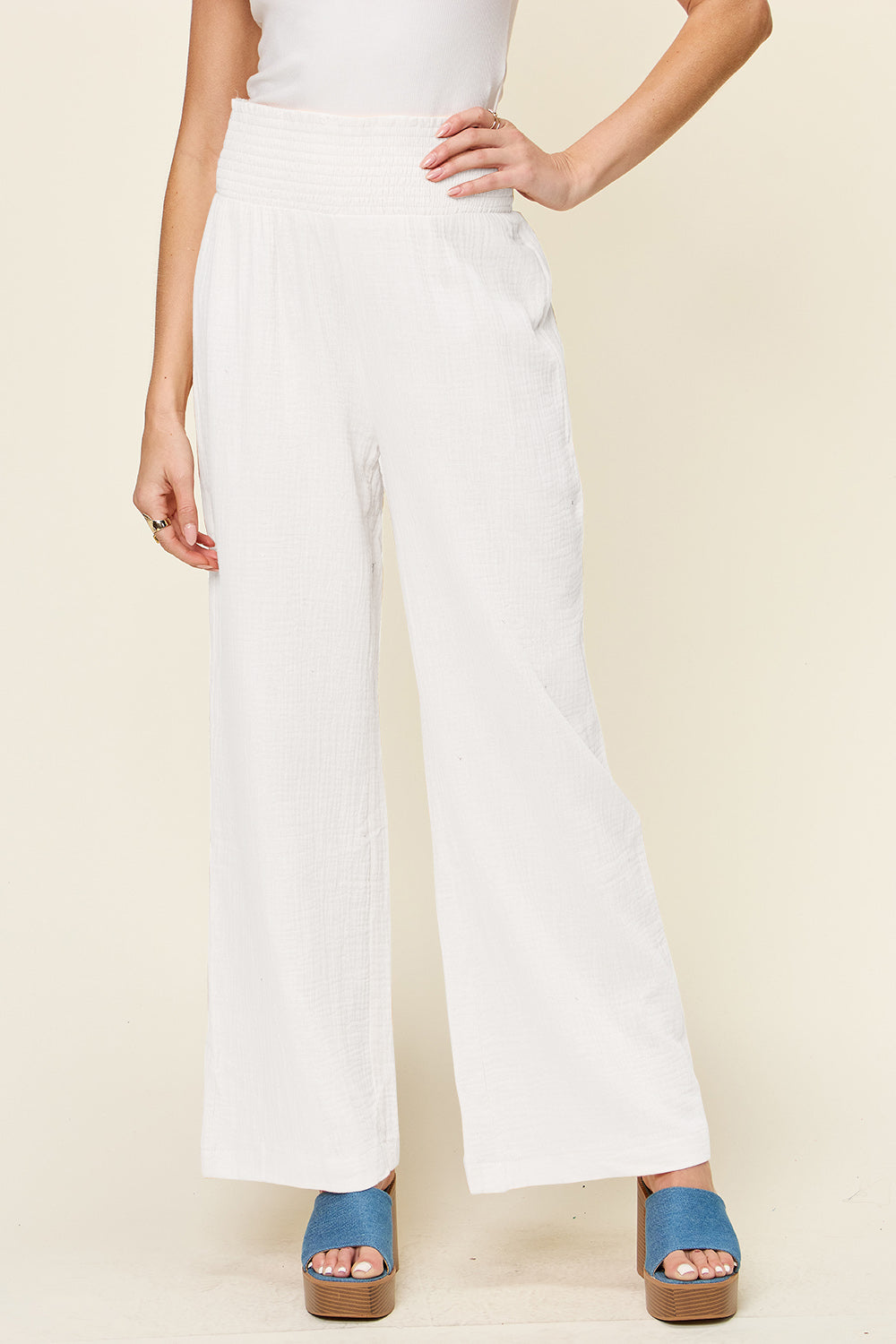 A person wearing Double Take Full Size Texture Smocked Waist Wide Leg Pants in black and a white 100% cotton top is standing with one hand on their hip. They are also wearing blue open-toe sandals.