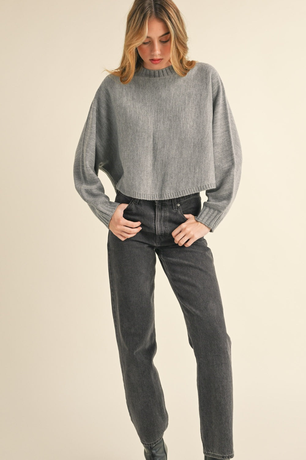 A person with long, light brown hair is facing away, wearing a Mable Round Neck Dolman Sleeve Cropped Sweater in grey paired with dark jeans, against a neutral background.