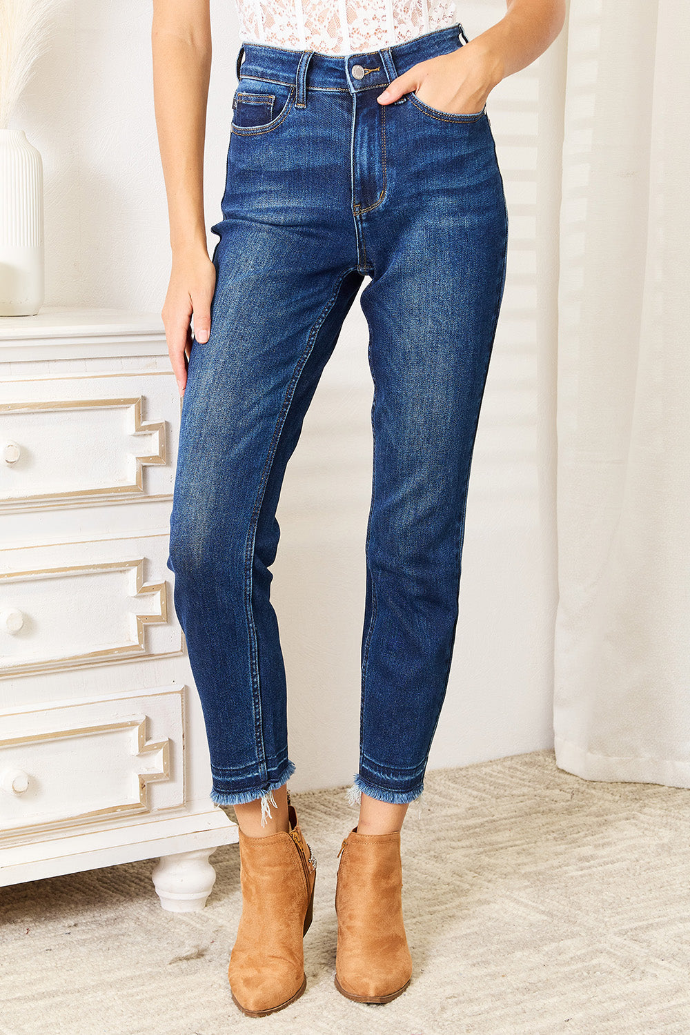 A person wearing Judy Blue Full Size High Waist Released Hem Slit Jeans in indigo wash and a white cropped top stands by a white dresser, showcasing a flattering fit.