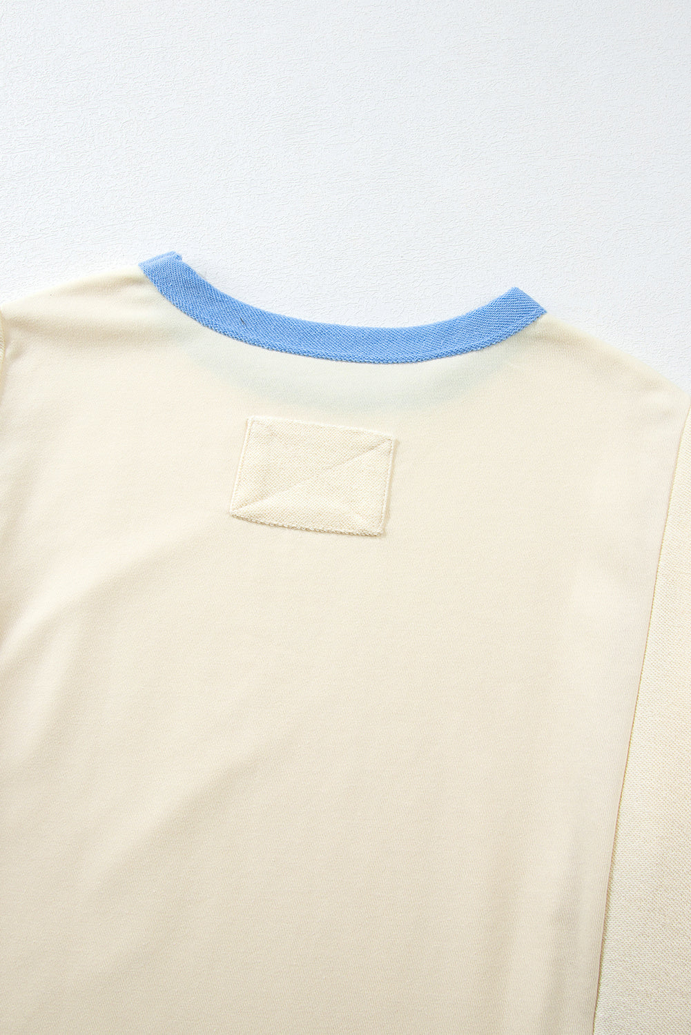 A person with long, wavy, light brown hair stands facing away, wearing the Beige Star Patchwork Exposed Seam Oversized Sweatshirt paired with denim shorts. The background is a plain, light peach wall, adding an edgy vibe to the look.