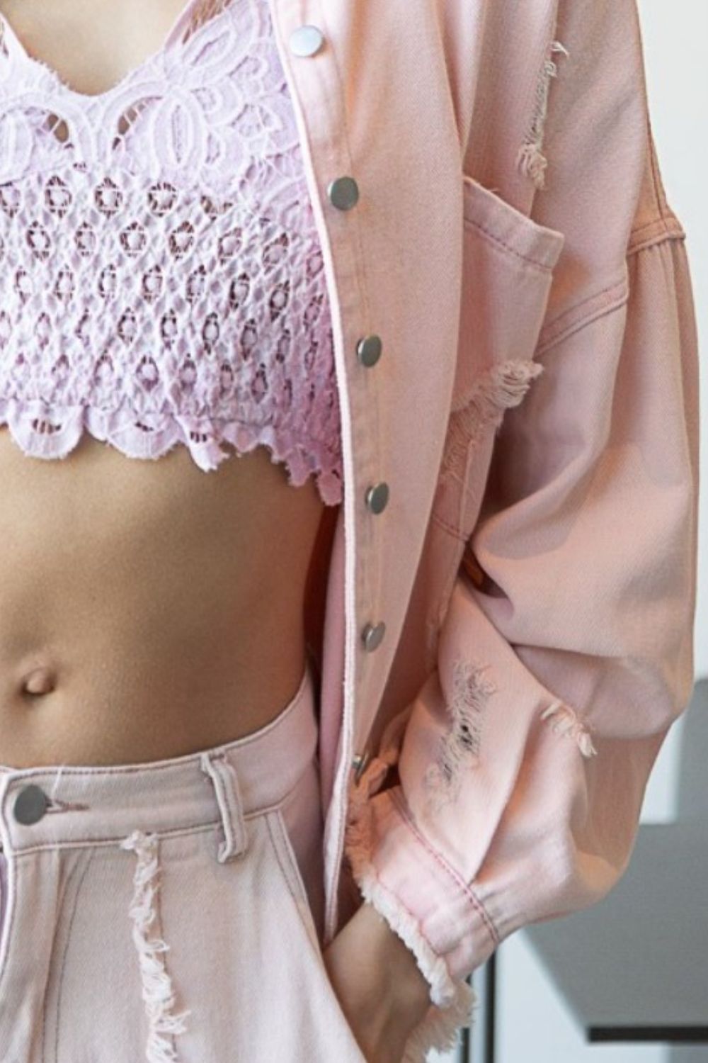 A woman wearing a POL Button Down Raw Hem Distressed Shacket in pale pink over a lavender lace crop top and matching pale pink distressed pants, standing indoors near a window.