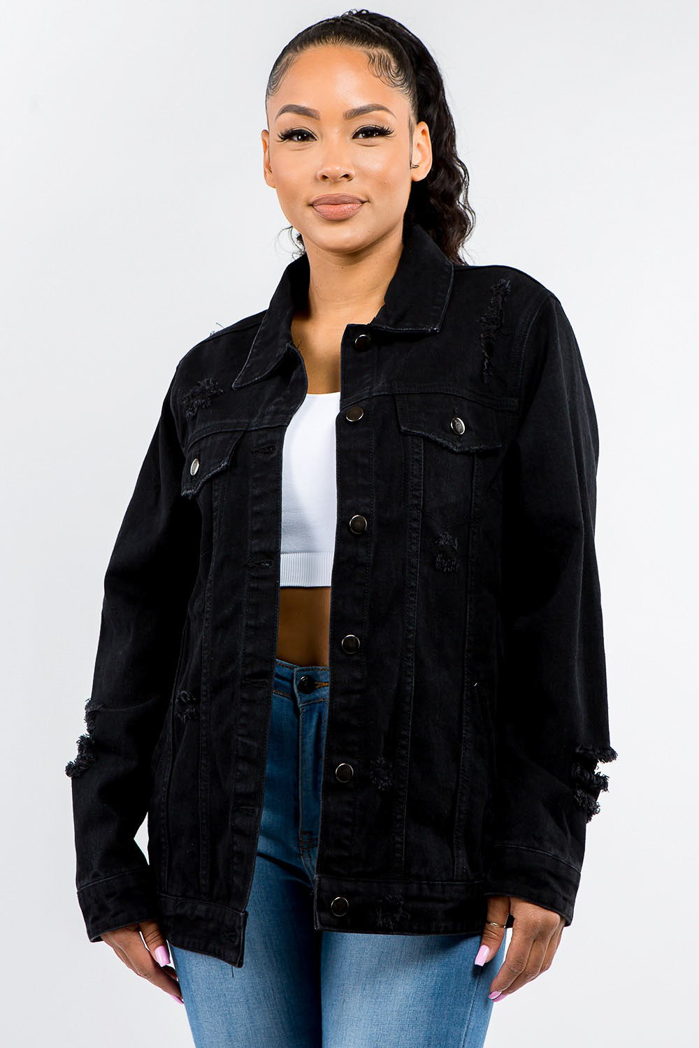 A woman stands against a plain background, epitomizing casual outfits with a touch of vintage look, wearing the American Bazi Full Size Button Up Distressed Denim Jacket over a white crop top and jeans.