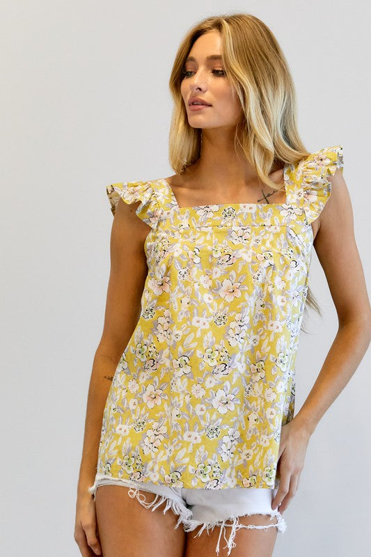 A woman in a vibrant summer wardrobe stands against a plain background, wearing the Floral Printed Ruffle Sleeveless Top paired with white shorts, with one hand casually tucked in her pocket.