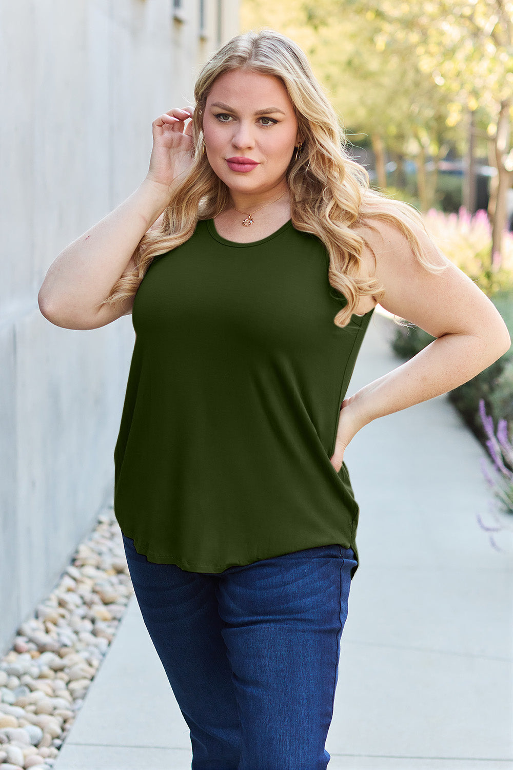 A woman wearing sunglasses, blue jeans, and the Basic Bae Full Size Round Neck Curved Hem Tank is standing in front of a concrete wall, holding her glasses with one hand and carrying a beige shoulder bag. Her basic style complements the slightly stretchy fabric of her outfit, ensuring comfort while maintaining chic simplicity.