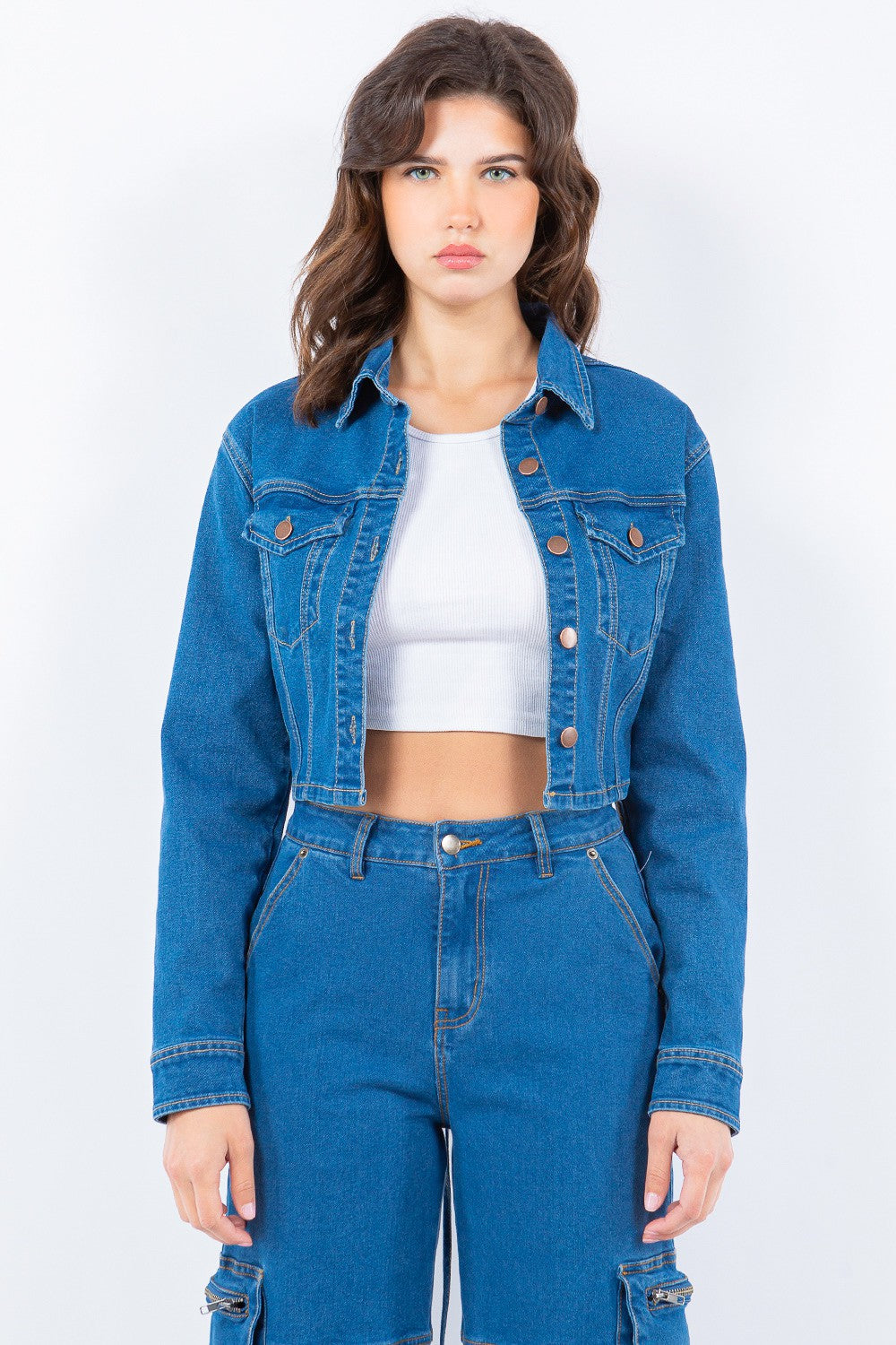 A person with long hair is seen against a plain background, wearing an American Bazi Laced Back Cropped Denim Jacket in blue denim, a white crop top, and matching blue denim pants.