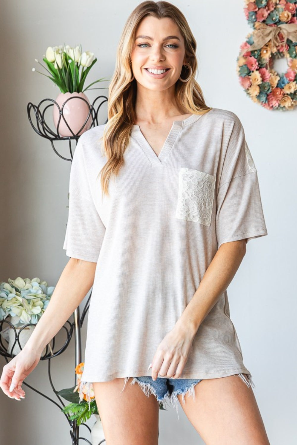 A woman wearing the Heimish Full Size Lace Front Pocket Drop Shoulder Top stands indoors near a decorative metal stand adorned with flowers.