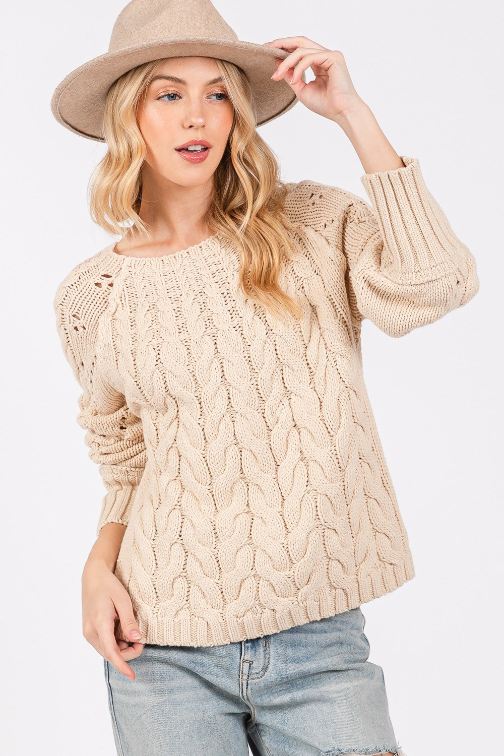 A person wearing a cozy, oversized SAGE + FIG Cable-Knit Long Sleeve Sweater in beige, light blue jeans, and a matching beige hat.