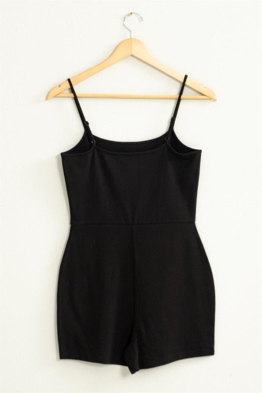 The Loving Me Fitted Sleeveless Romper, featuring a fitted bodice and spaghetti straps in black, hangs gracefully on a wooden hanger against a white wall.